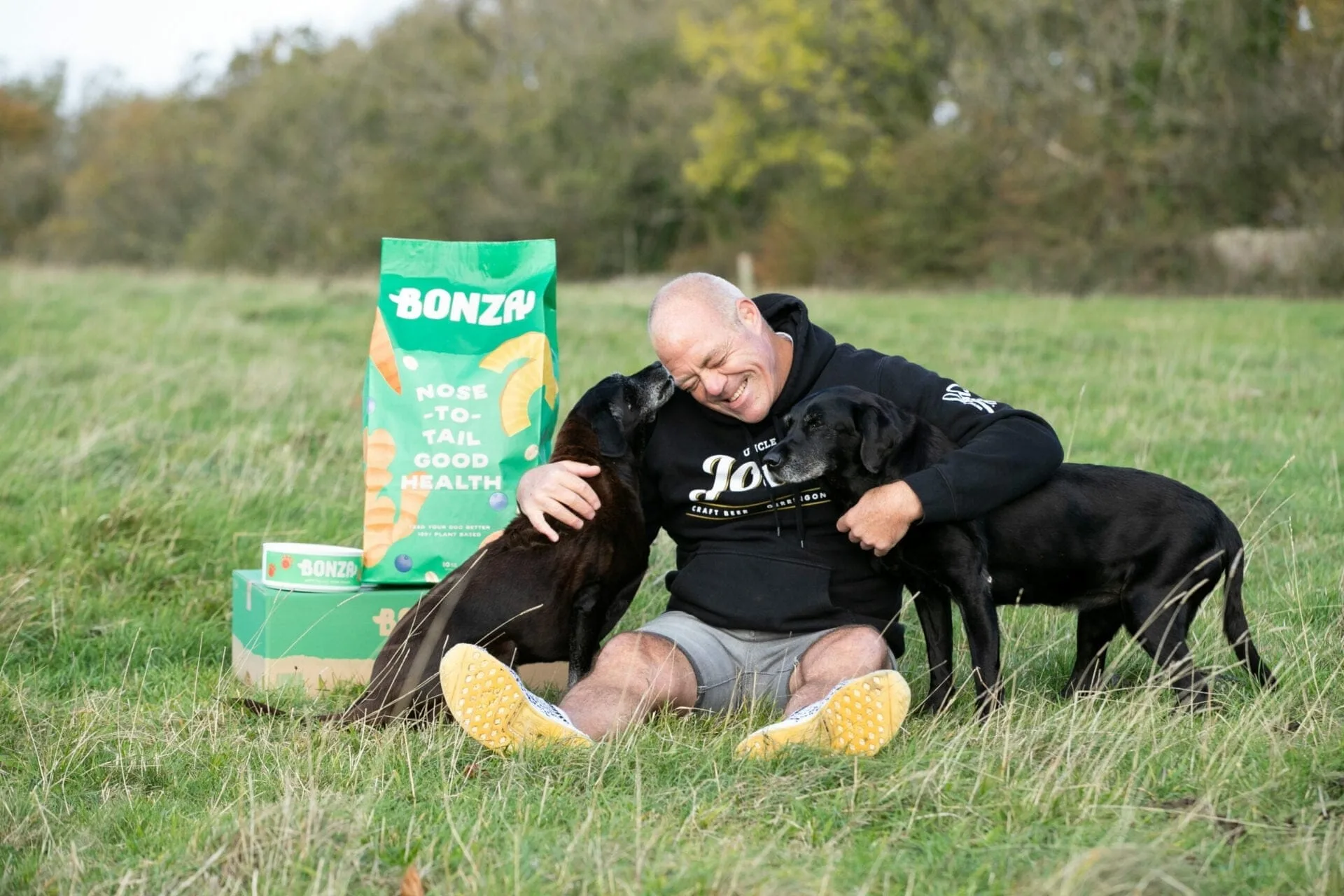 Bonza founder Glendon Lloyd sitting in a field with his two labradors, beside a bag of Bonza plant-based dog food